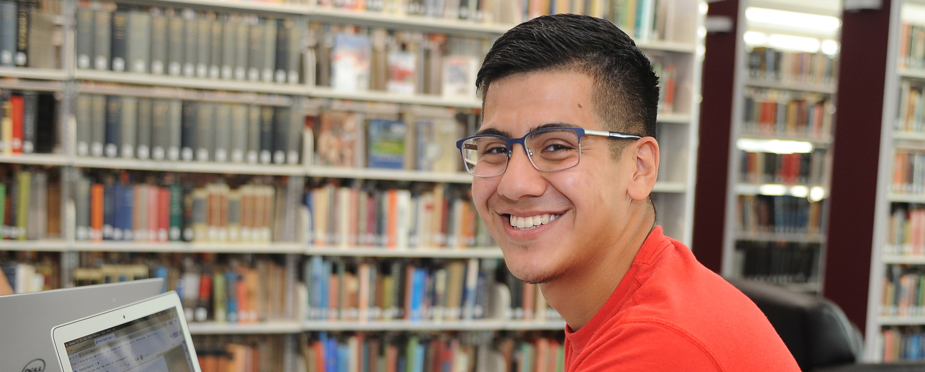Male student in library with shelves of books in the background. He is seated in front of a computer screen but smiling at the camera.