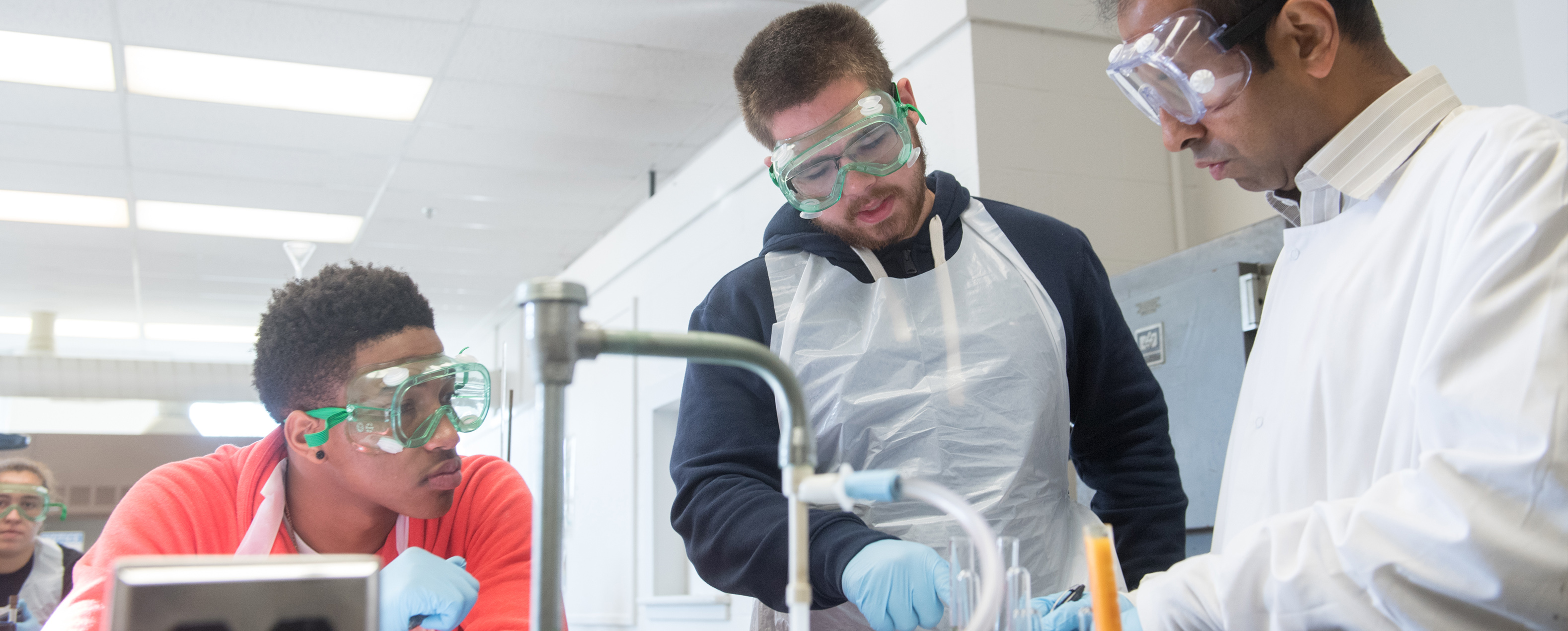 Two young males at a lab station working with test tubes and faculty member Dr. Dutta. All wearing safety goggles.