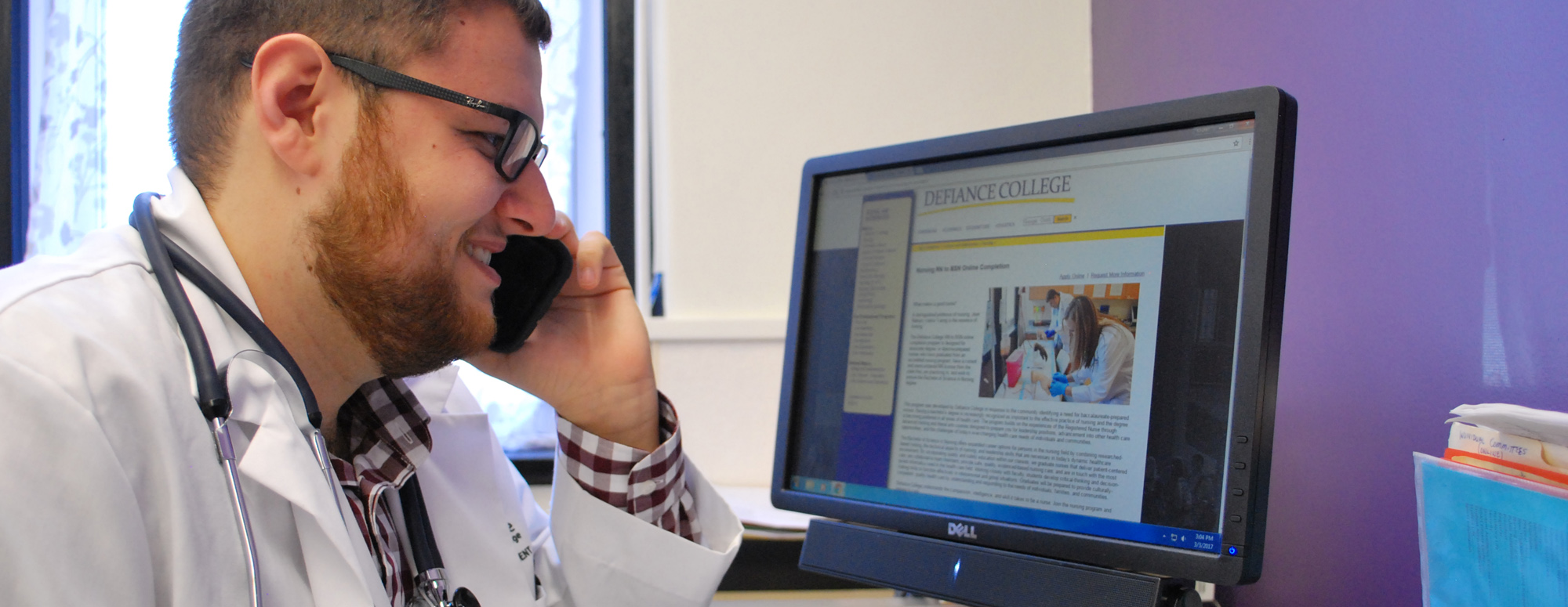 male student in white nurse jacket talking on telephone while sitting at a desk and looking at a computer screen that is displaying the Defiance College nursing webpage.