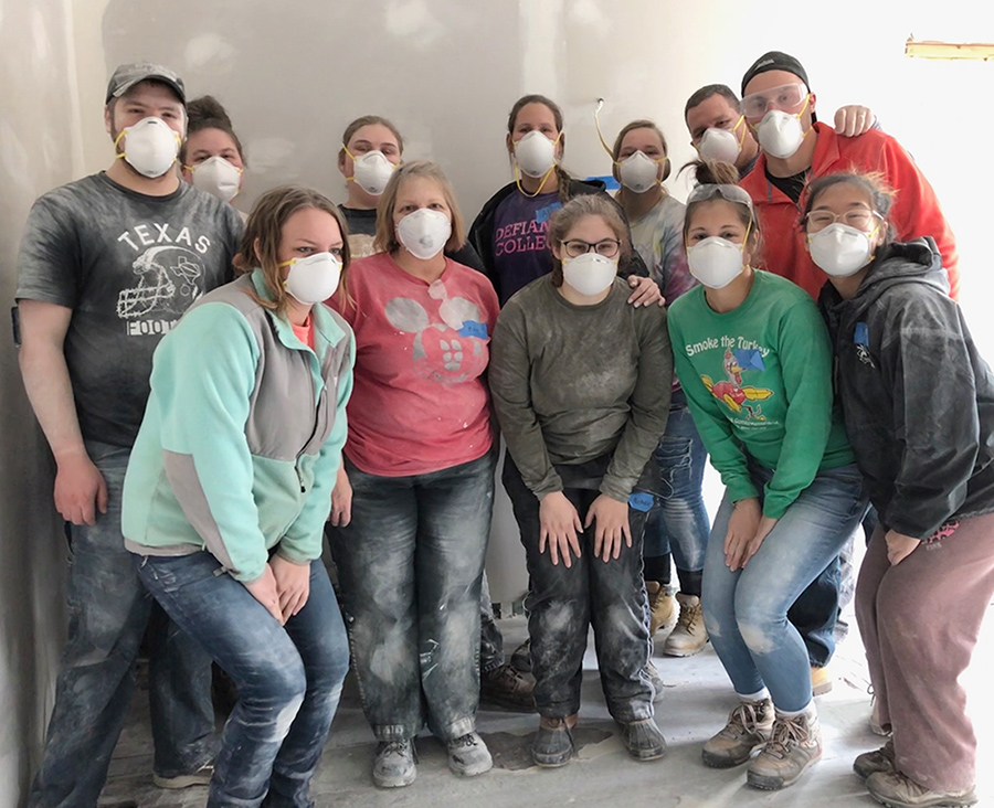 12 individuals standing in a line facing the camera, wearing white construction face masks. Their clothes are dirty from working on a house.