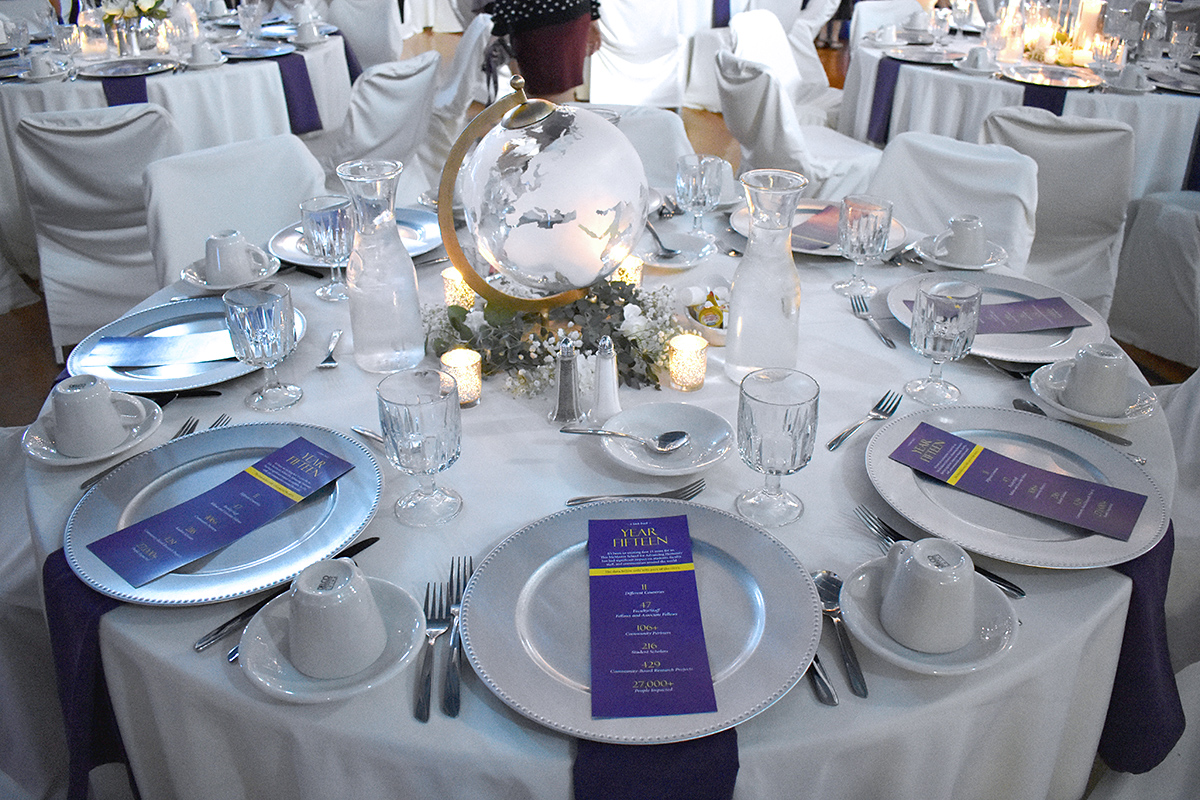 Table set up with white table cloth, purple banners, plates, glasses, silverware, and centerpiece.