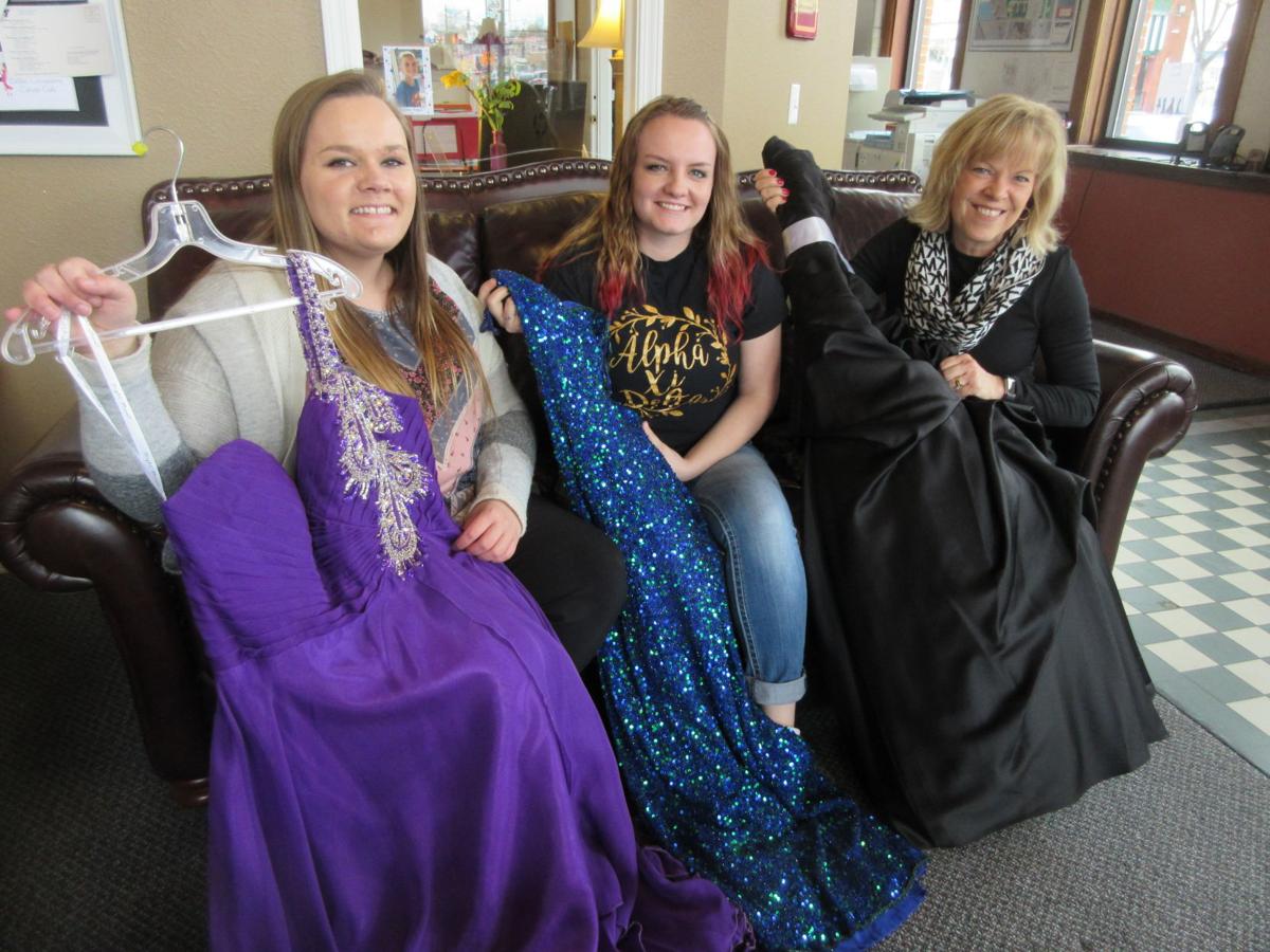 Two female students with dark blonde hair and one blonde woman sitting on a leather couch and holding up dresses that are purple, blue, and black.