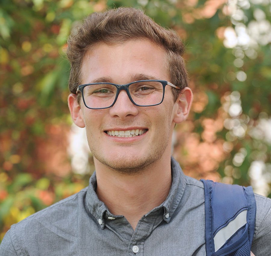 Young man with light brown hair wearing glasses. He is smiling at the camera. Shades of green from leaves are blurry in the background.