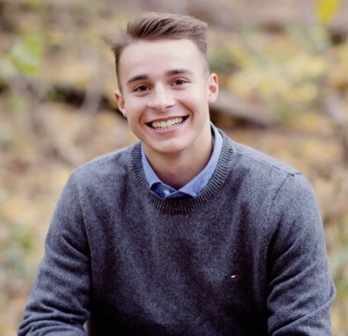 Young man in a blue collared shirt with a grey sweater over it. He is smiling at the camera.