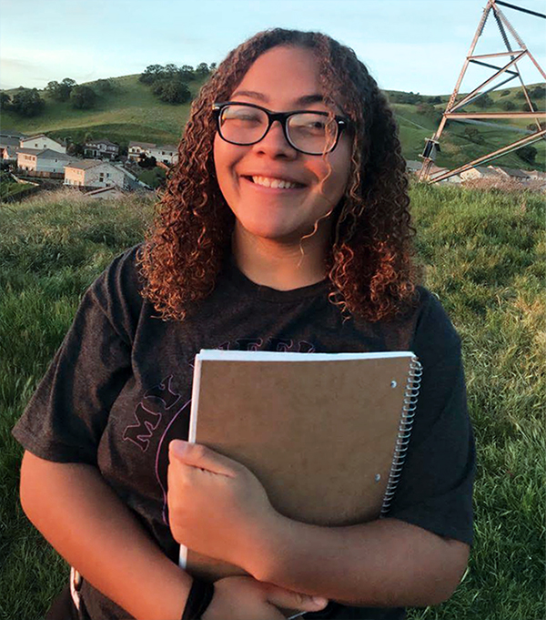 Young woman with shoulder length dark brown hair and black glasses. She is in a dark grey shirt, holding a spiral notebook to her chest, and smiling at the camera.