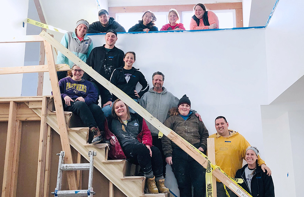 13 people standing on and above a stair case that is under construction. They are wearing hoodies and jackets of all colors and smiling at the camera.