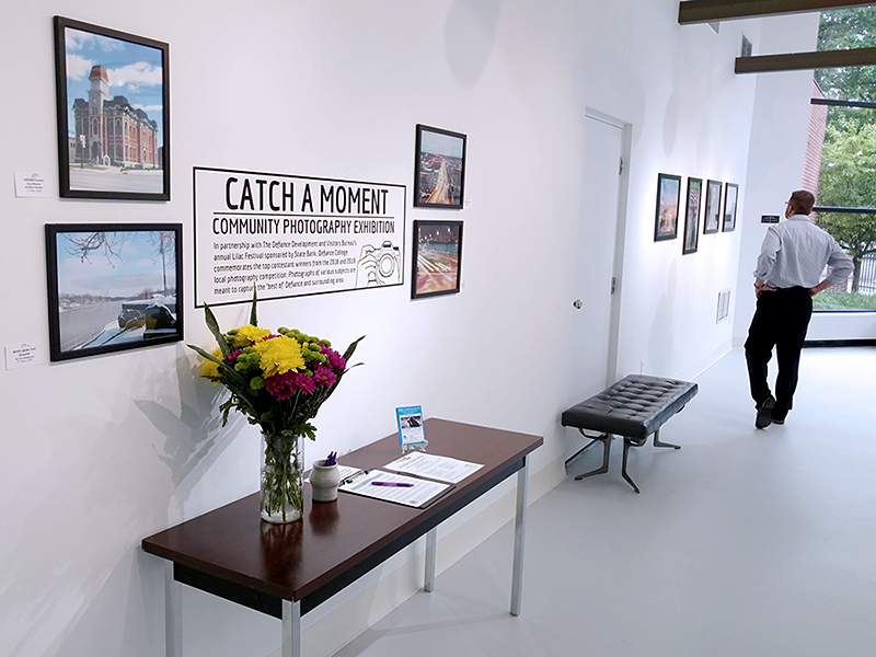 Large photos of scenes in the city of Defiance are hanging on the gallery wall. A table with flowers and a guestbook are sitting against the wall.