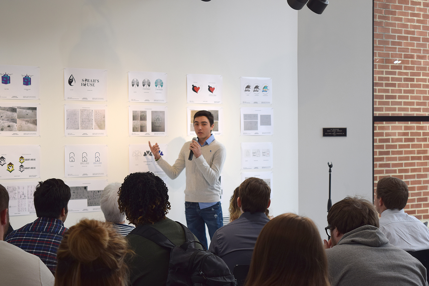 Student Federico Gutierrez holding a microphone and speaking in front of an audience who is watching him point at his design on the exhibit wall.