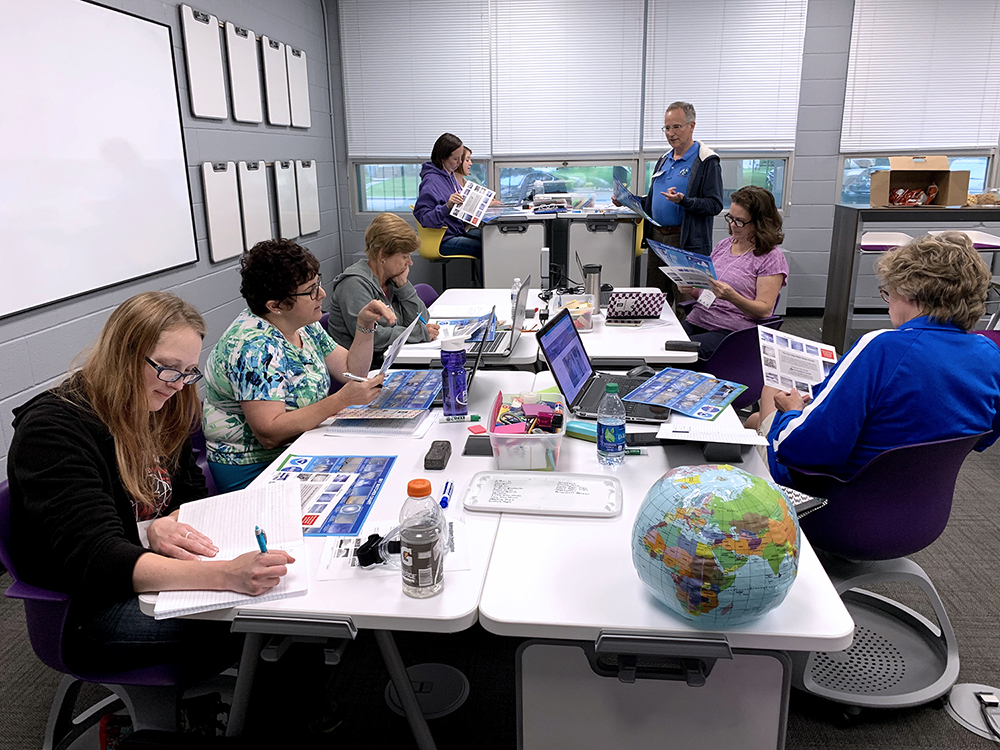 Seven teachers sitting around tables with books, notebooks, and laptops. A man is standing in the back, speaking to them.