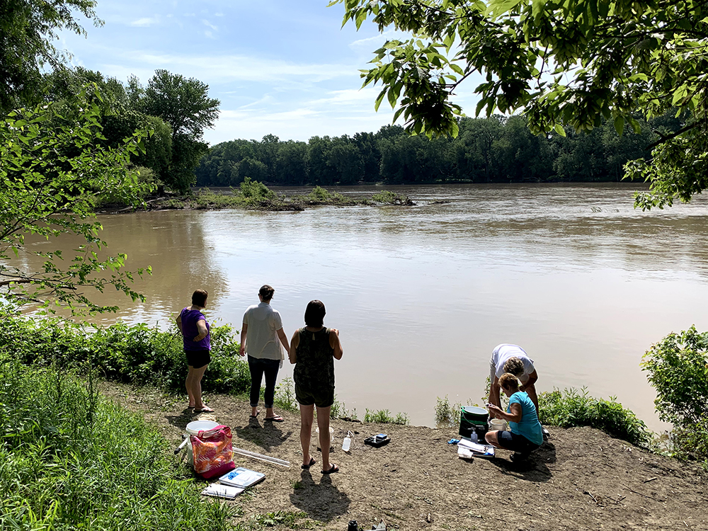 Five women standing near the Maumee river bank with equipment to test the water.