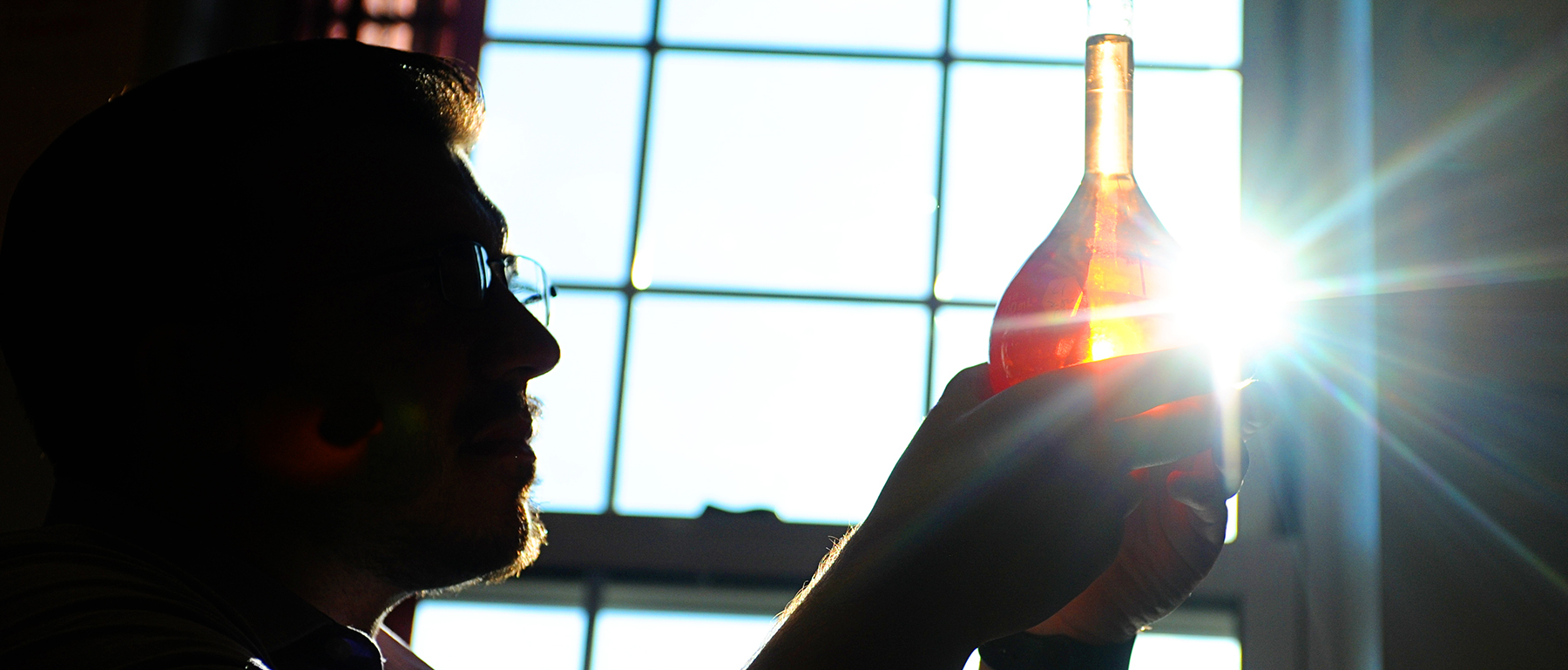 A male student stands in front of a window holding a vile of liquid as the sun shines behind him.