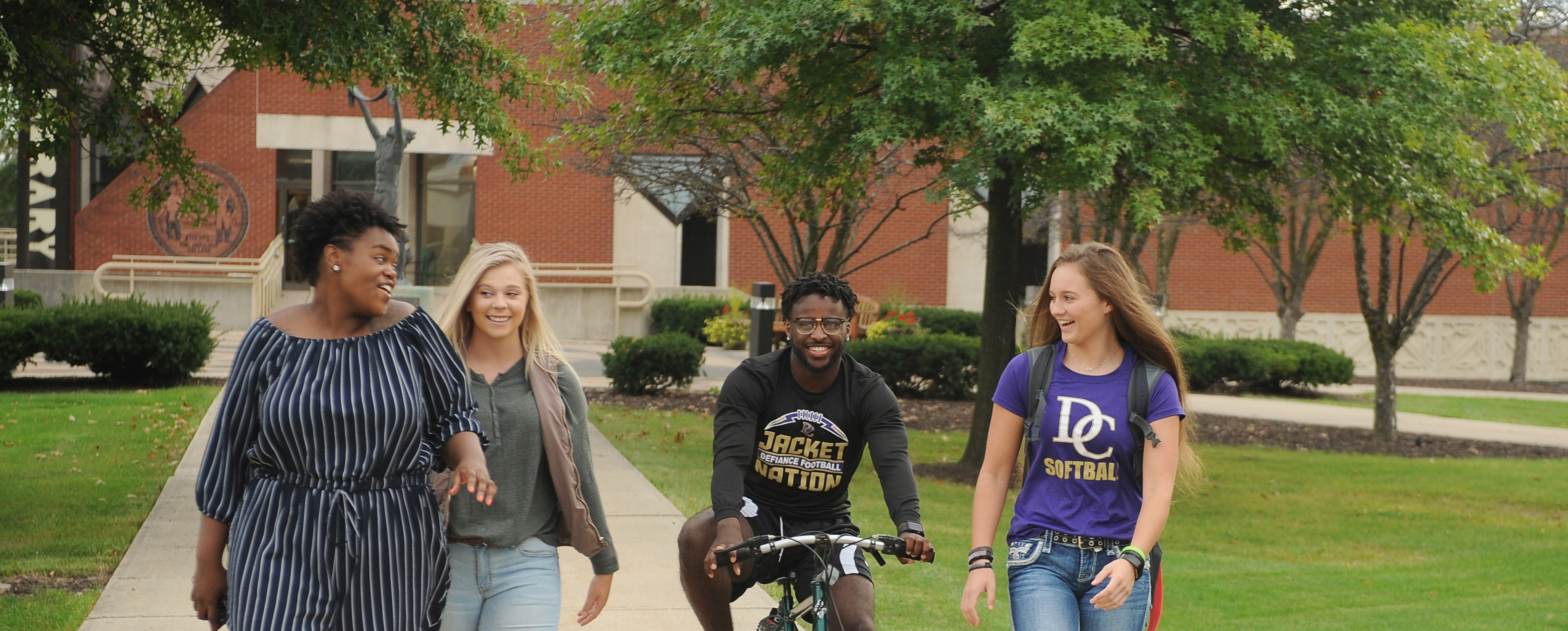 Three female students walking on a campus sidewalk with a male student on a bike. All are actively talking with each other and smiling.