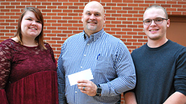 female student in red shirt and male student in black sweater smiling at the camera. between them is a man in a blue checkered dress shirt holding a check.