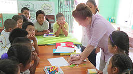 Bright room with mint green walls covered in learning posters. Young woman in lilac shirt writing at a desk surrounded by 17 children and 1 other young woman helper.