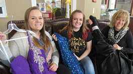 Two female students with dark blonde hair and one blonde woman sitting on a leather couch and holding up dresses that are purple, blue, and black.