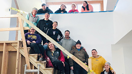 13 people standing on and above a stair case that is under construction. They are wearing hoodies and jackets of all colors and smiling at the camera.