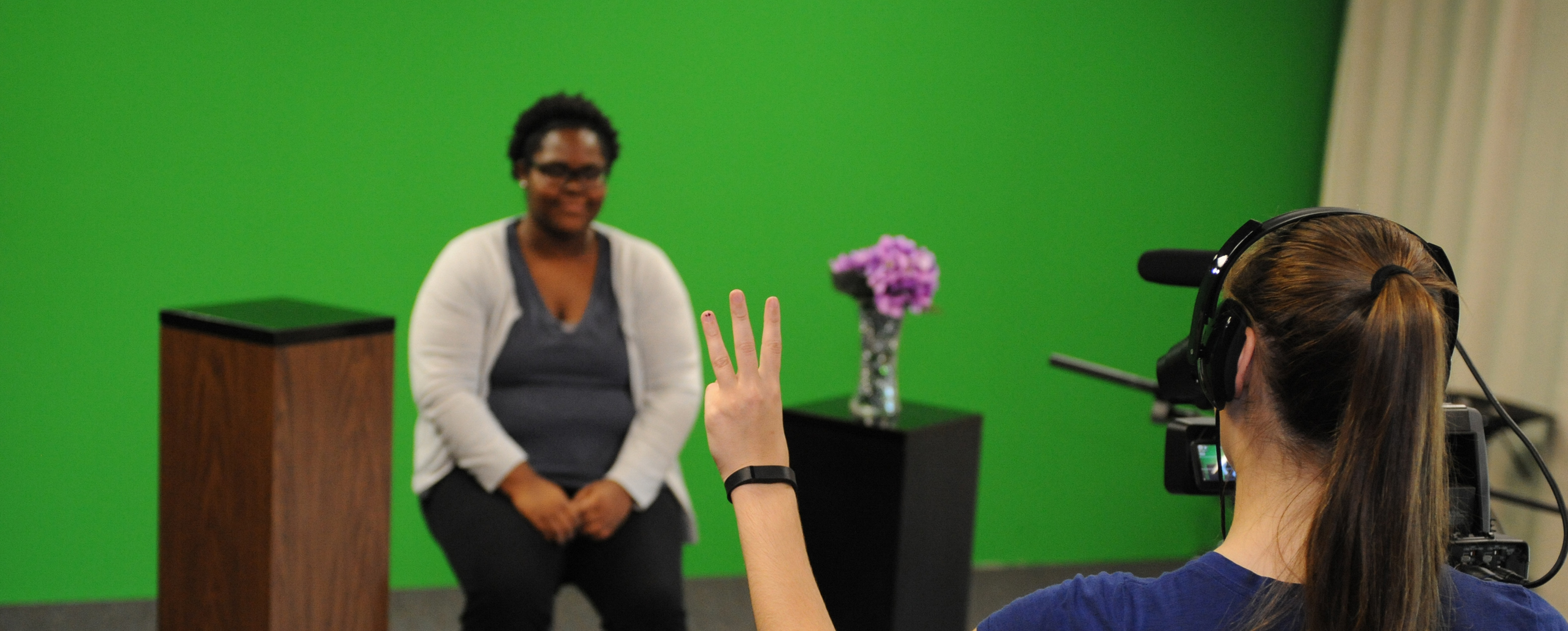 Student sitting in the background in front of a green screen. Another student is in the foreground, directing a camera at the first student and holding up three fingers in a countdown position.