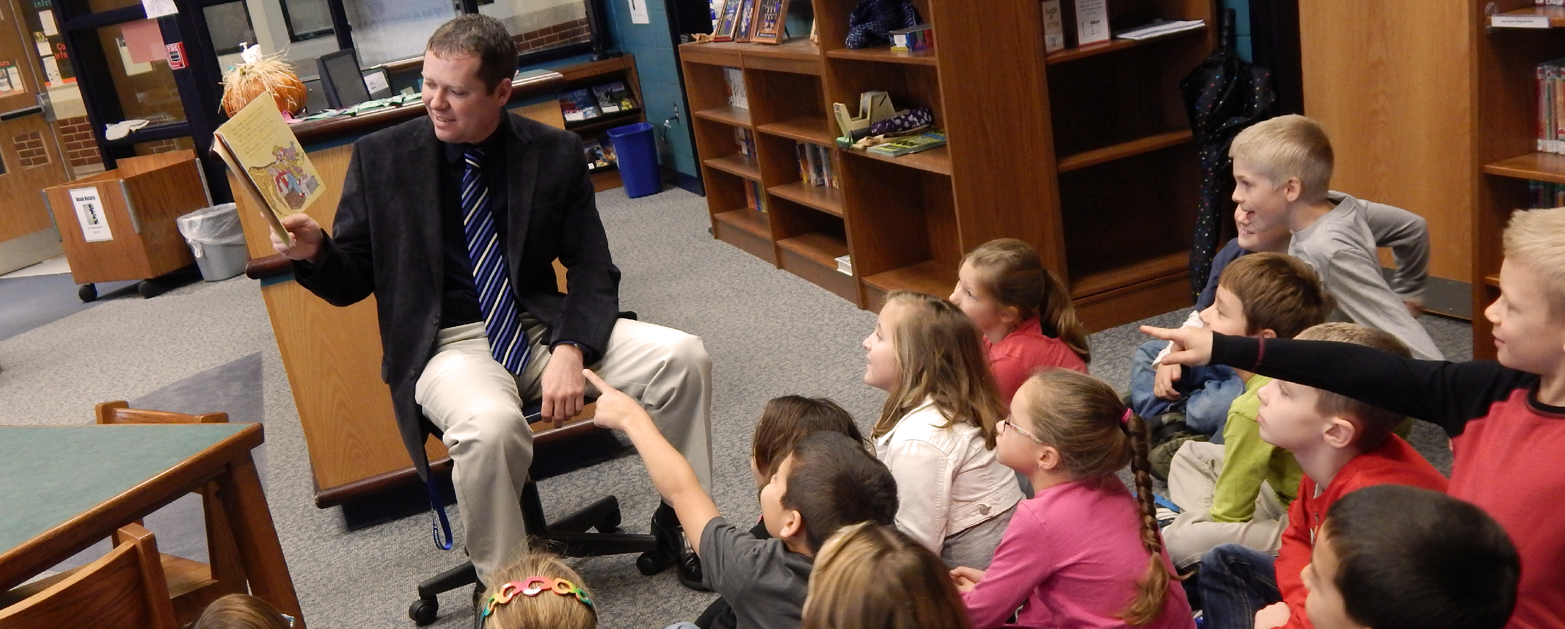 Man in business attire sitting on a chair with a book in front of a row of children who are pointing at the book and watching him share the book with excited expressions on their faces.