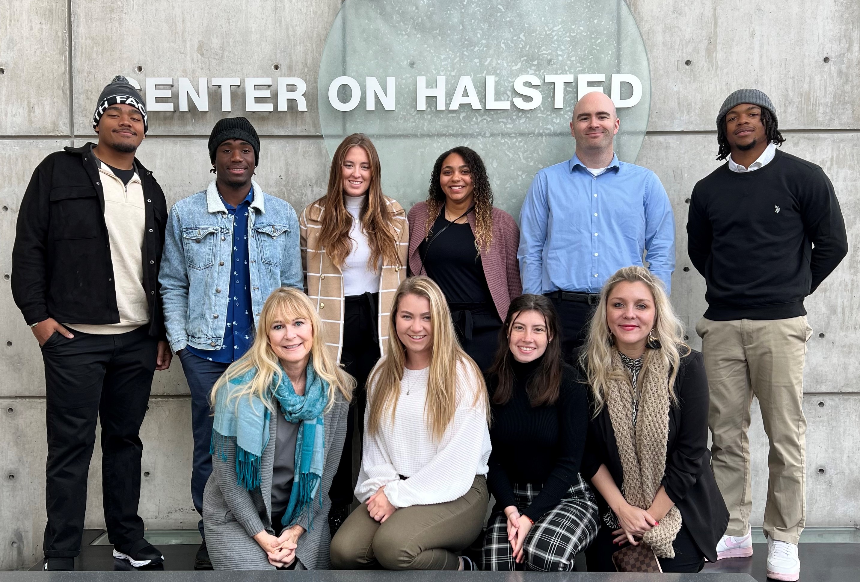 10 individuals from Defiance College smiling in front of a grey conrete wall. The words Center on Halsted are on the wall.