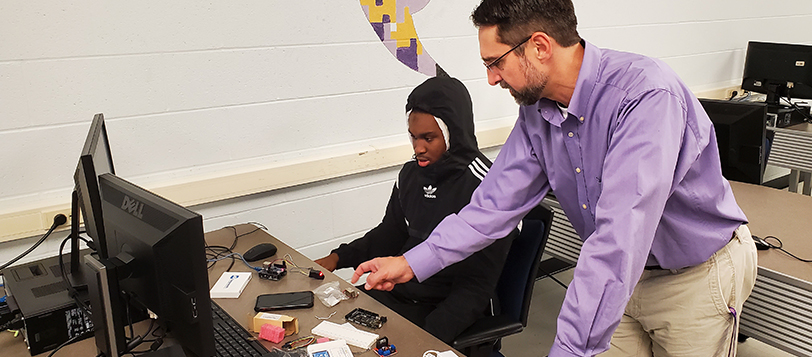 A professor teaching a student about computer science by a computer. There's a phone and computer parts on the desk.