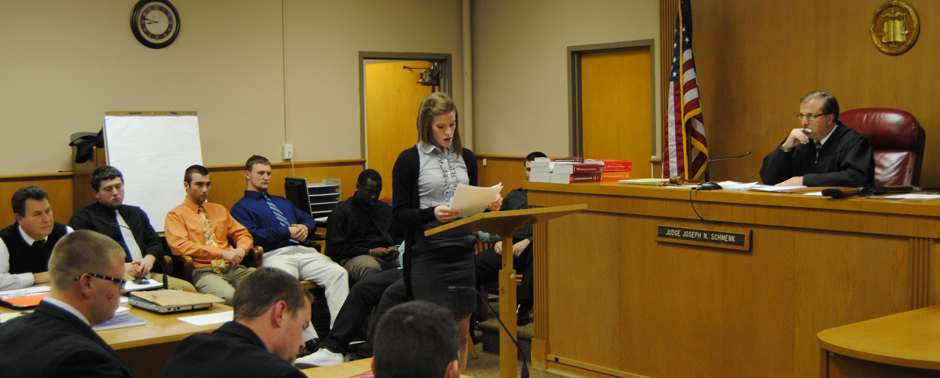 Nine men sitting around the corner edge of a court room. A young blonde woman in nice clothes is standing at a podium reading off papers while a judge listens from behind his desk.