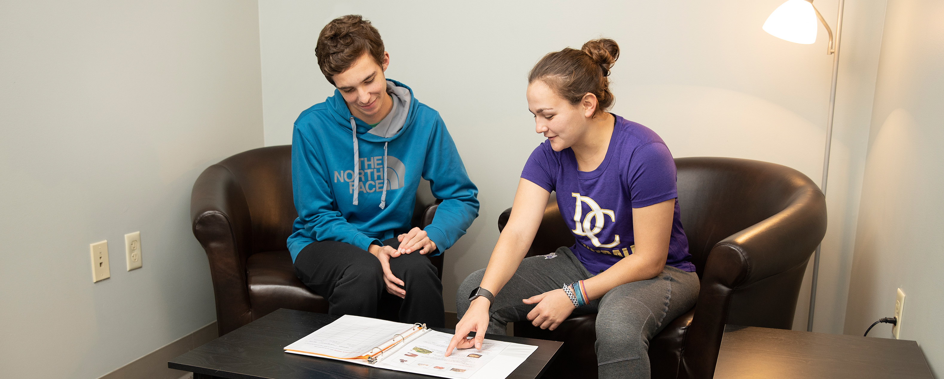 Male and female student sitting on black chairs and looking at a binder on a table.