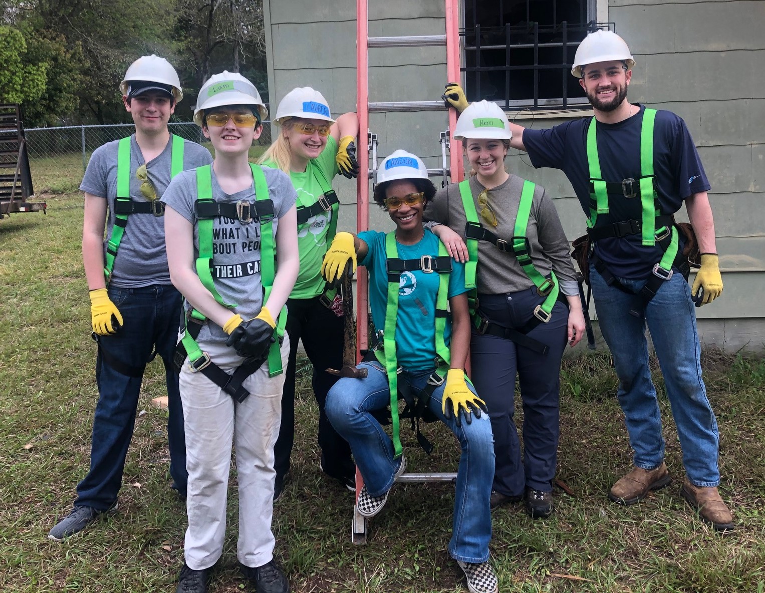 six students dressed in work gear and hard hats standing in front of a construction site