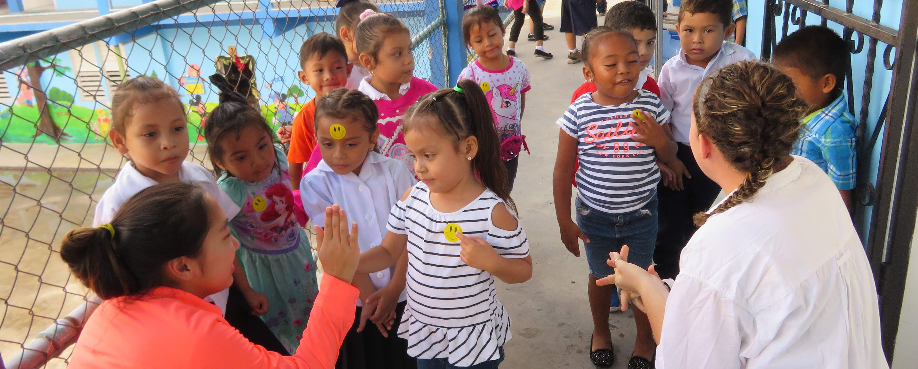 Two older girls have their backs to the camera, facing a group of 12 small children.