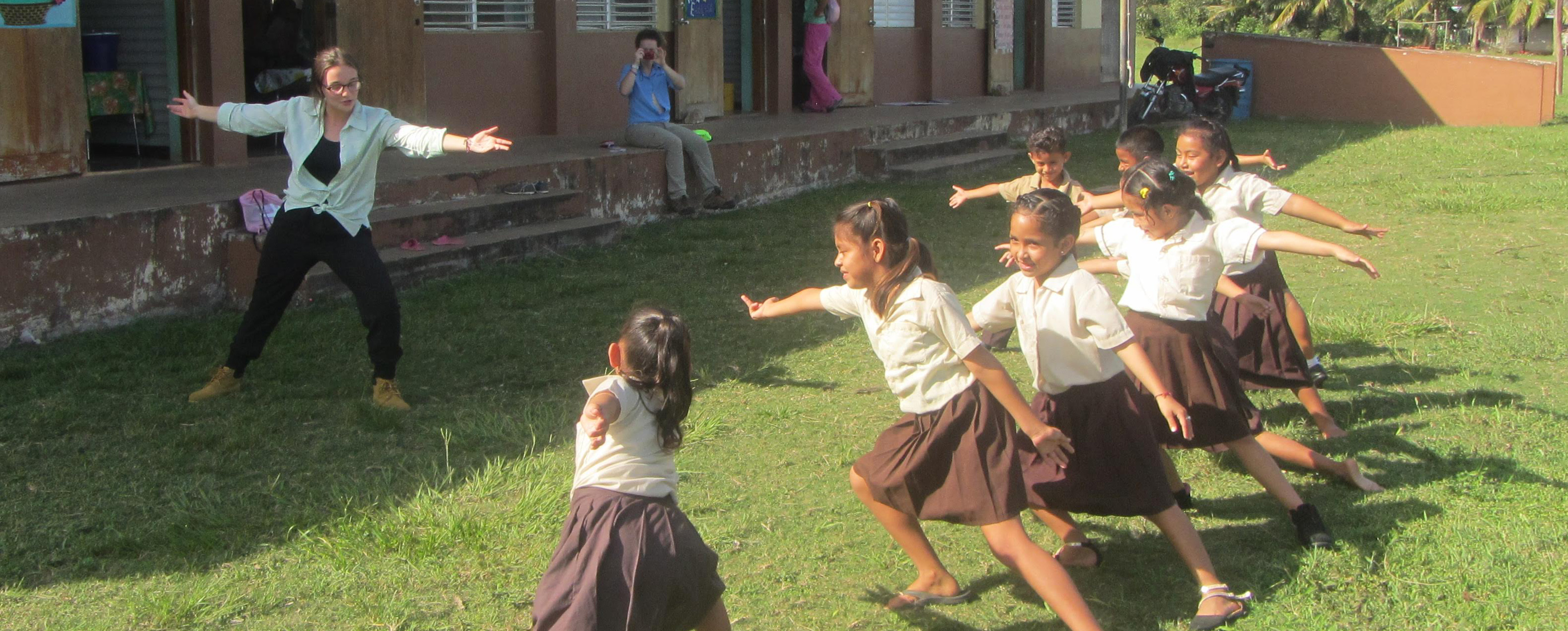 Female student with arms out stretched, standing in front of seven children who are copying her pose. They are outdoors in the grass, in front of a tan and brown building.