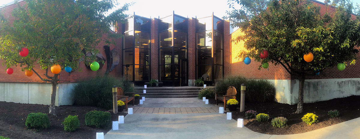 Outdoors with trees, dark brown mulch, and bushes framing a sidewalk leading stairs up to a brick building with tall glass windows.