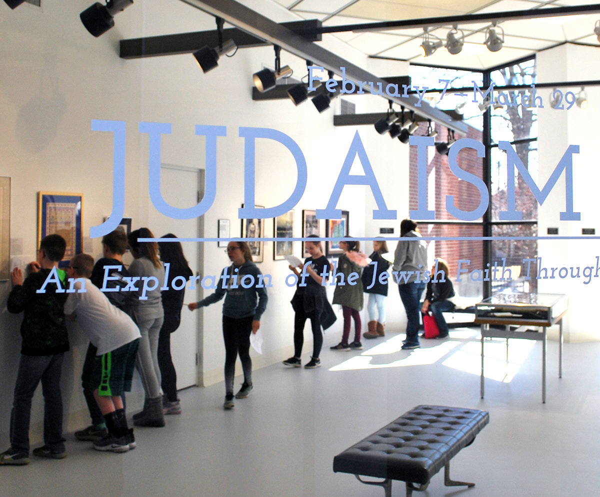 View through the window at a group of people looking at framed works hanging on the gallery wall. The window has a decal that says February 7 - March 29, Judaism, An Exploration of the Jewish Faith