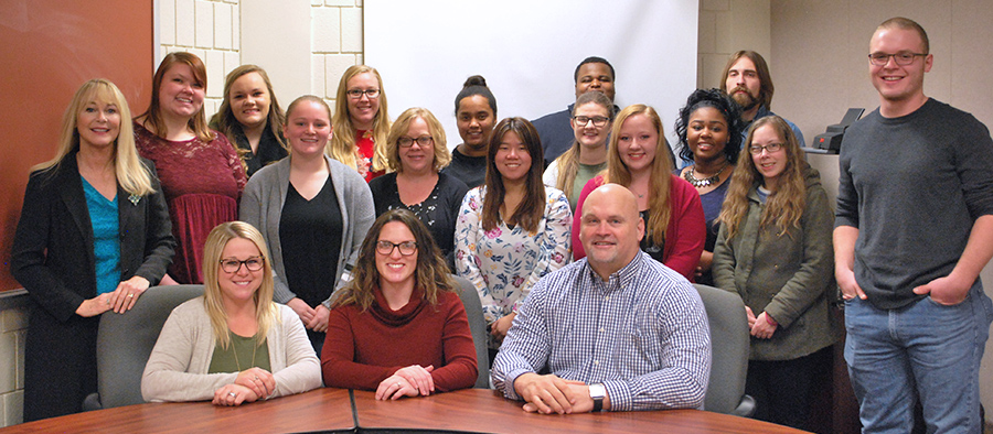 14 women and 5 men gathered in front of a conference table, smiling at the camera