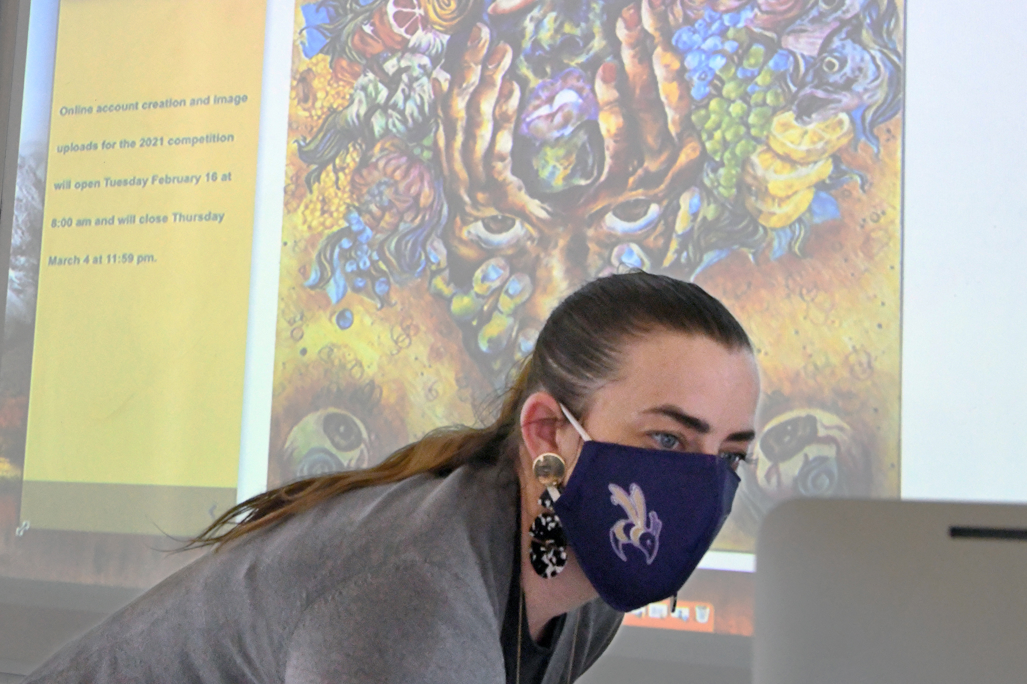 Woman with a long pony tail wearing a mask in front of a projected screen with artwork on it
