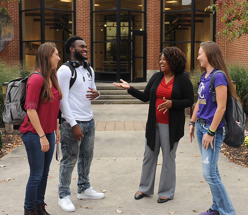 Two female students and one male student outside of a brick building with tall glass windows talking and laughing with Mercedes Clay