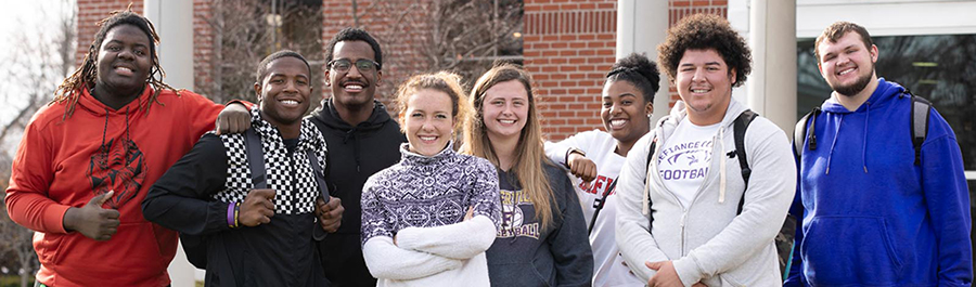 Nine students standing before the brick front of Hubbard Hall on campus, taking a selfie with a phone and all smiling at the camera.