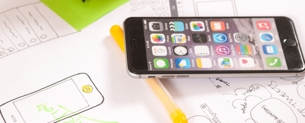 A smartphone with colorful app icons lies on top of hand-drawn sketches of mobile app designs, next to a yellow pen.