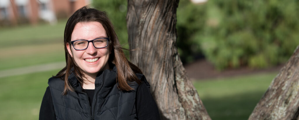 A young woman with straight brown hair, glasses, and a black vest smiles while standing outside near a tree, with green grass and blurred buildings in the background.
