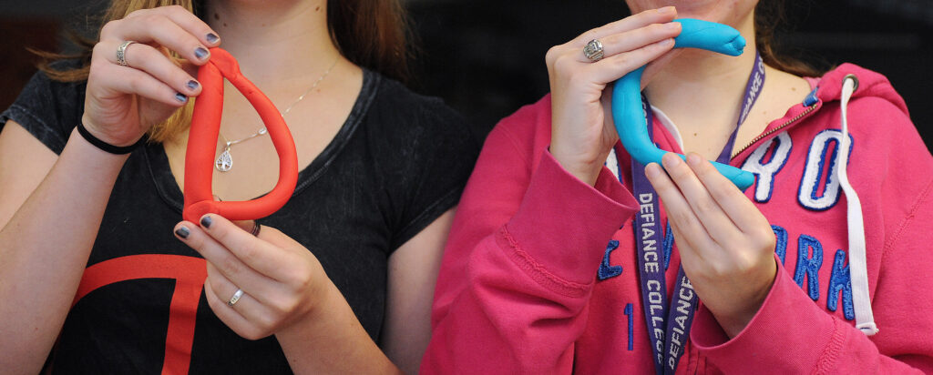 Two people hold colorful clay shapes, one red and one blue, resembling the Greek letters delta and theta. Their faces are not visible; only their hands and upper bodies are shown.