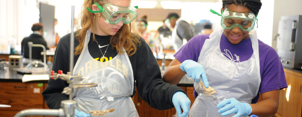 Two students wearing safety goggles, gloves, and aprons work together on a lab experiment in a science classroom, handling materials and focusing on their project. Other students are visible in the background.