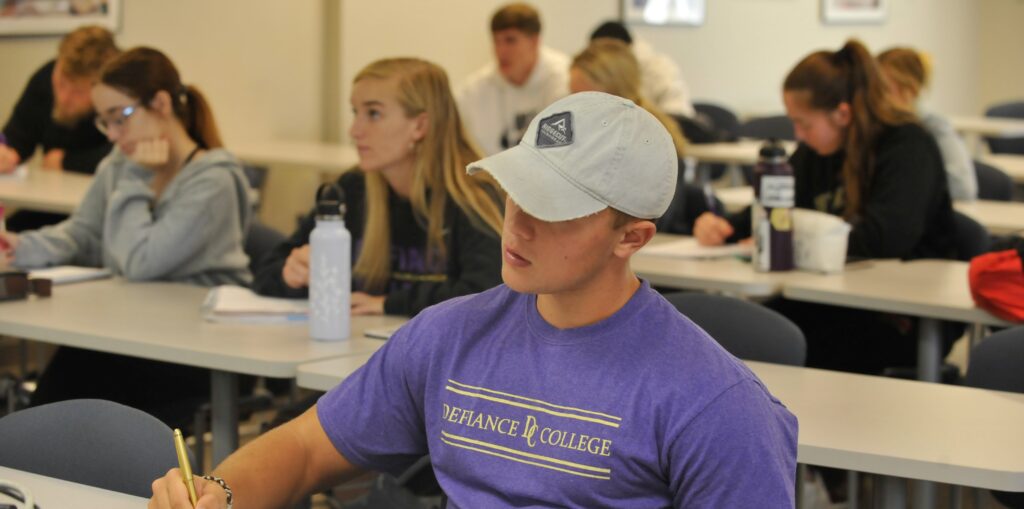 College students sit at desks in a classroom, taking notes and listening. A student in the foreground wears a light gray cap and a purple Defiance College shirt, focused on writing. Other students are blurred in the background.