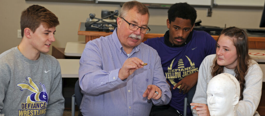 A teacher explains while holding a small object, sitting with three students in Defiance wrestling and Defiance Yellow Jackets shirts, next to a white mannequin head in a classroom setting.