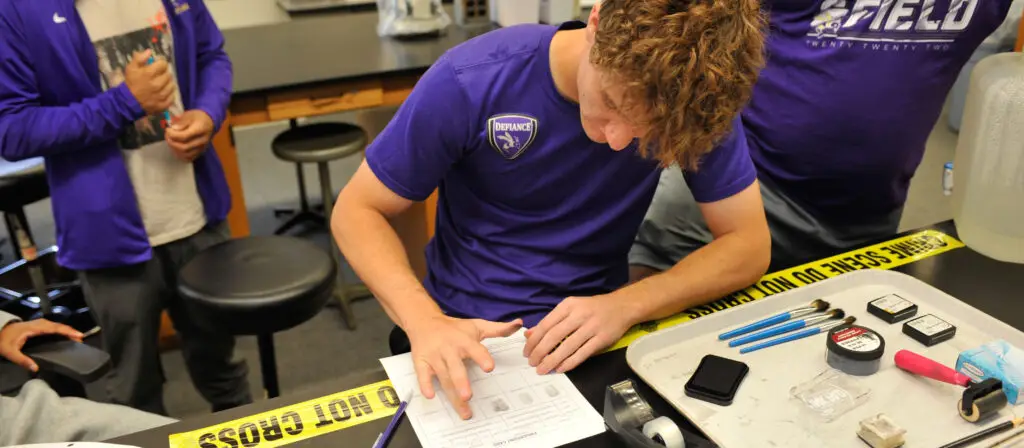 A student in a purple shirt examines a worksheet at a lab table, surrounded by fingerprinting tools and evidence tape, while other students stand nearby.