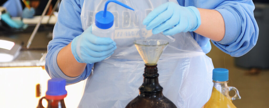 A person wearing blue gloves and a plastic apron pours liquid into a funnel placed on a brown bottle, surrounded by other bottles and lab equipment.