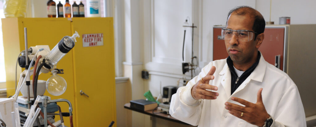 A scientist in a white lab coat and safety goggles gestures with his hands while talking in a laboratory, with lab equipment and chemical bottles visible in the background.