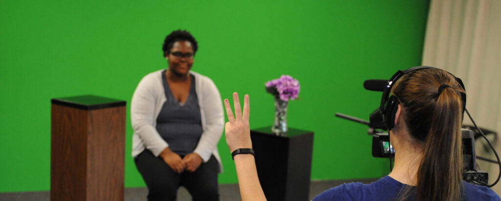 A woman sits on a stool in front of a green screen, with a podium and vase of purple flowers nearby, while another person behind a camera holds up three fingers to signal her.