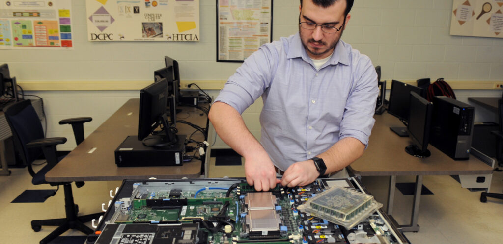 A man in a light shirt works on the internal components of a desktop computer in a classroom with multiple computers and posters on the walls.
