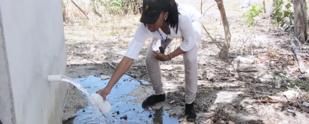A person in a white shirt and cap bends down to collect water from a pipe flowing out of a wall, filling a container in an outdoor, dry, and grassy area.