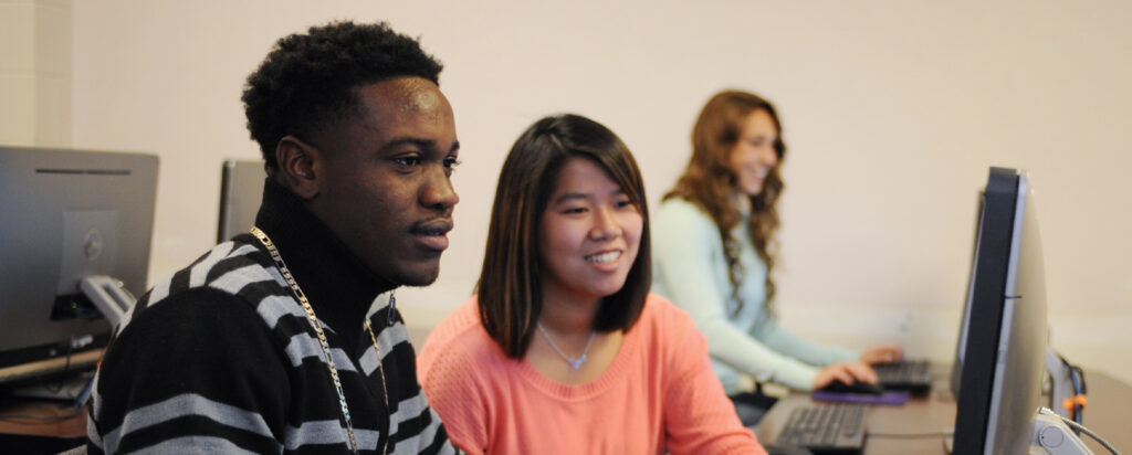 Two young adults, a man and a woman, sit together at a computer, smiling and focused on the screen. Another woman sits at a separate computer in the background, also smiling.