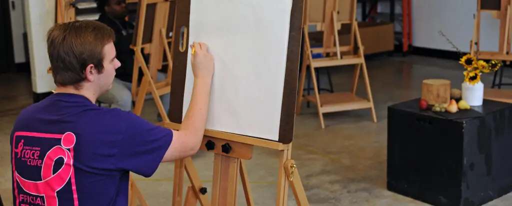 A person sits at an easel, drawing on a blank white canvas in an art studio. In the background are other easels and a table with a vase of sunflowers and some fruit set up for still life.