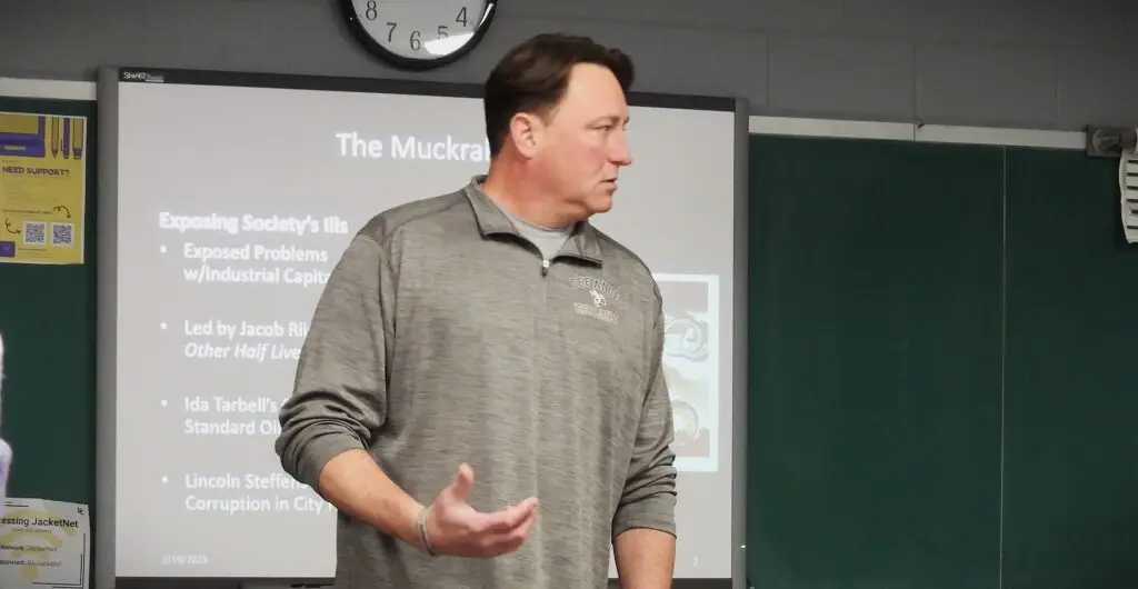 A man in a gray shirt stands and gestures while speaking in a classroom. Behind him is a projected presentation titled The Muckrakers, and a wall clock shows the time as 11:05.